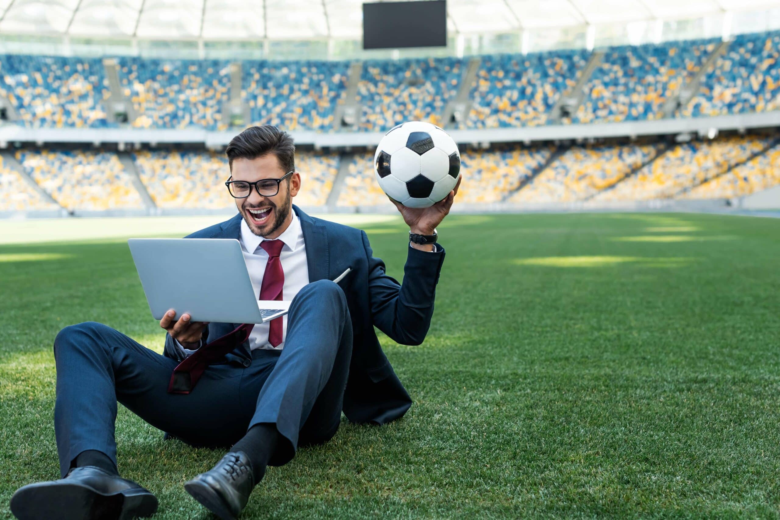 Arbitrázs jelentése a sportfogadásban - illusztráció a cikkhez. Egy öltönyös fiatalember laptoppal és focilabdával a kezében ül a stadion közepén és örvendezik, feltehetően a sportfogadási nyereménye láttán.